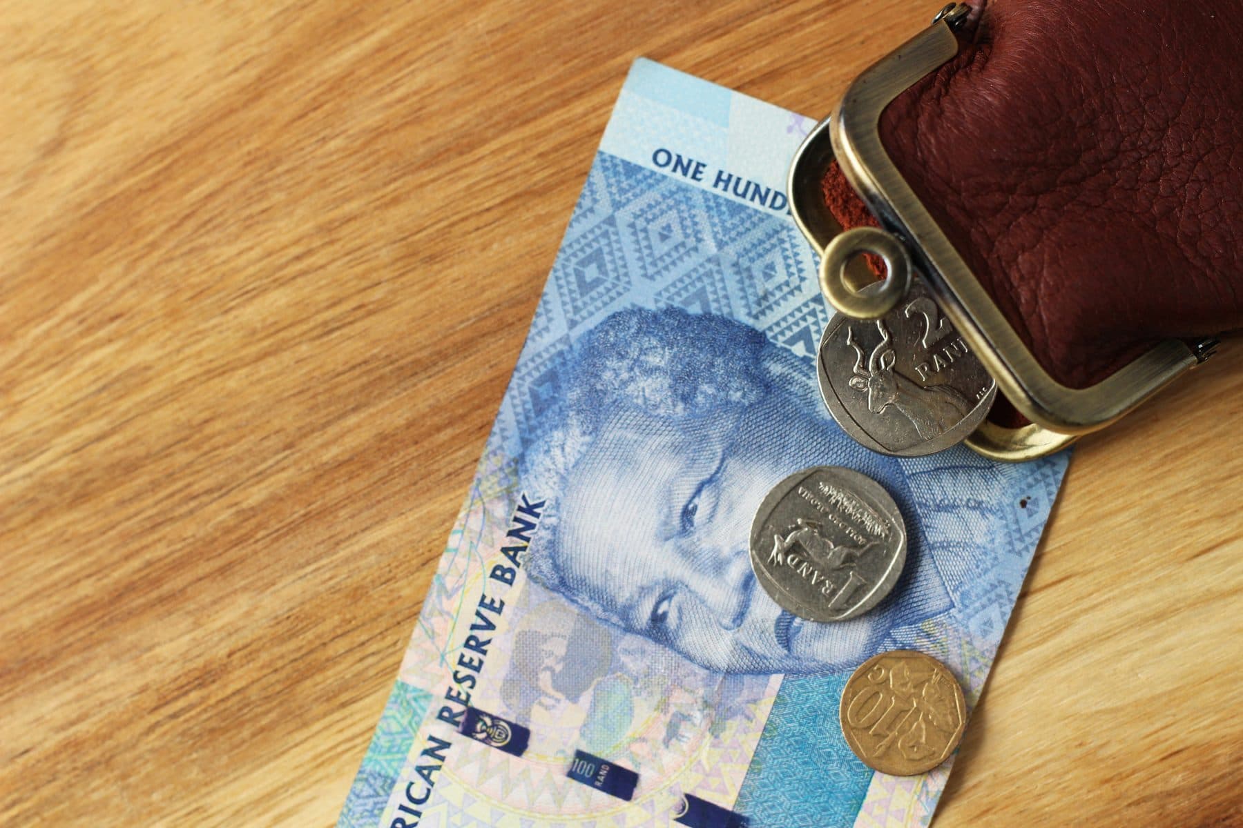 South African R100 note and coins spilling from a leather coin purse on a wooden surface